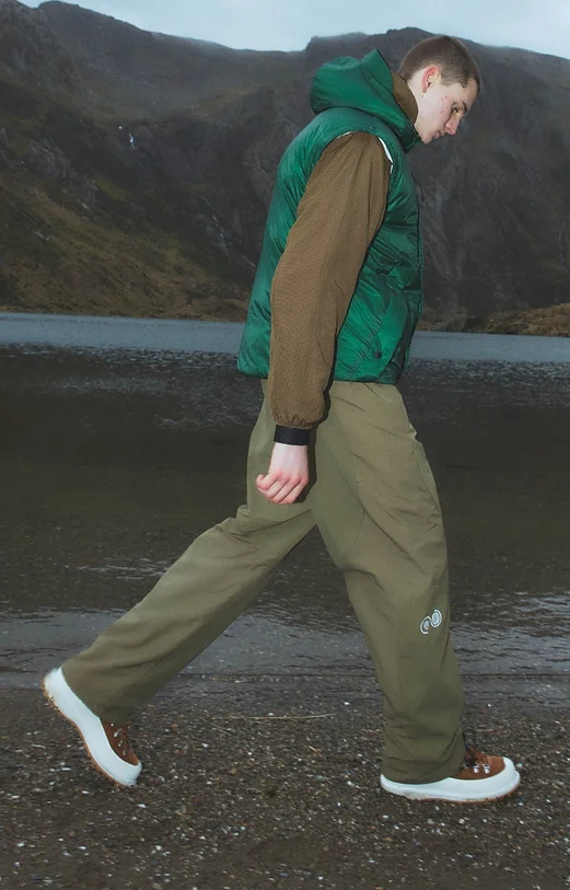 Man wearing a green quilted vest walking beside a lake and mountain