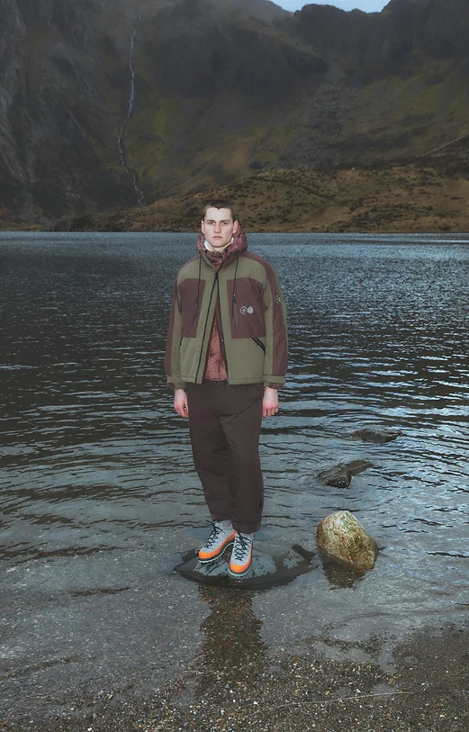 Man wearing a brown and green jacket, standing on a stone in a lake