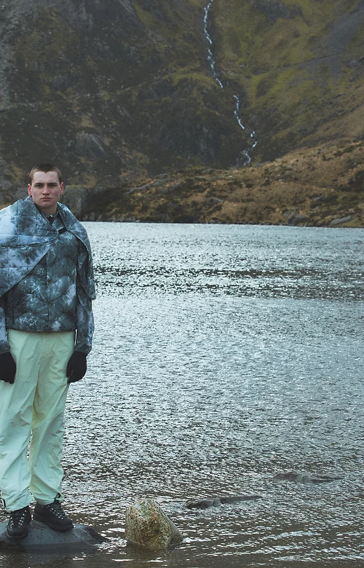 Man wearing a jacket with an "ice dye" pattern, standing on a stone in a lake