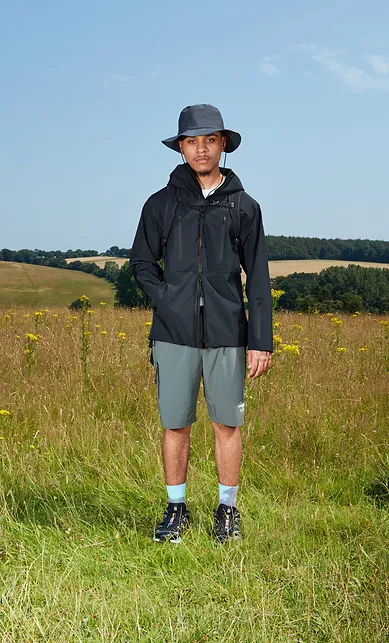 Man wearing a black jacket and grey shorts snd sun hat, standing in a field