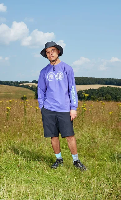 Man wearing a purple top, black shorts and a sun hat, standing in a field in the sun