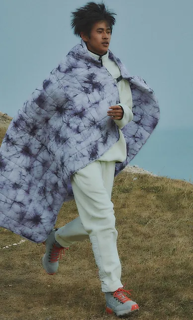Man wearing a quilted cape in ice dye pattern, walking along clifftops