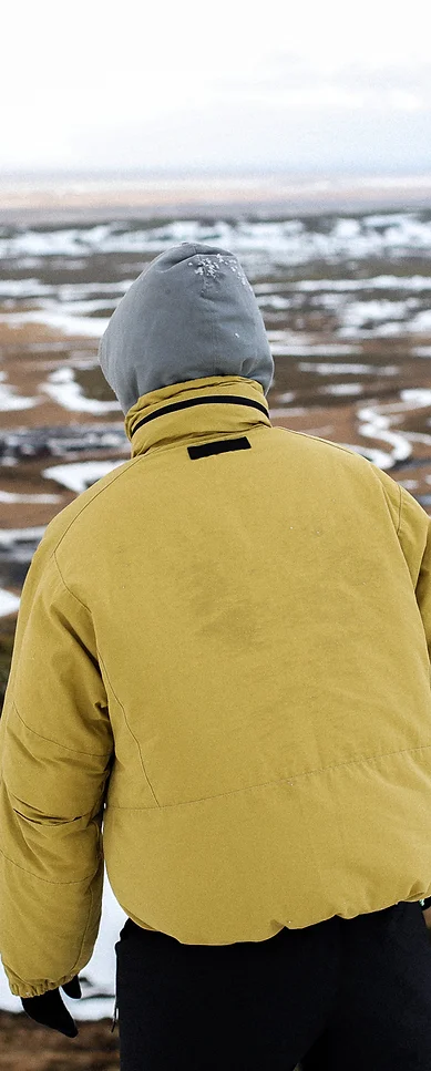 Man wearing a yellow jacket, black trouser and grey hood, walking through a snowy mountain landscape.